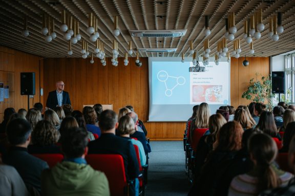 Ein Vortrag in einem Raum mit vielen Zuhörern und einer Präsentation im Hintergrund.