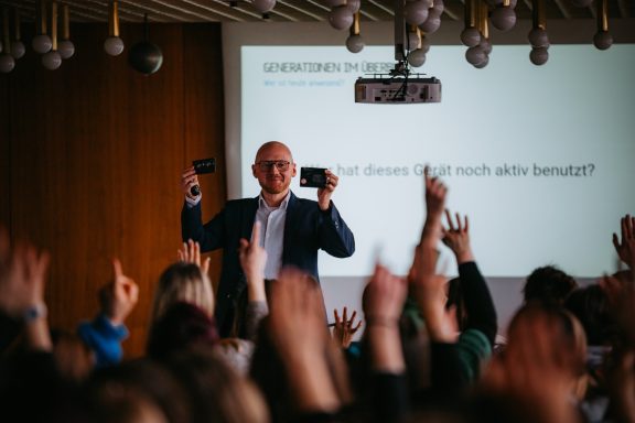 Ein lächelnder Mann hält zwei Handys hoch, während das Publikum die Hände hebt.