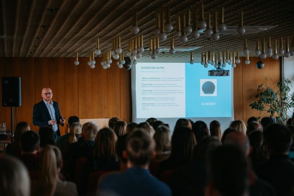Mann präsentiert vor Publikum in einem Konferenzraum mit Bildschirm im Hintergrund.